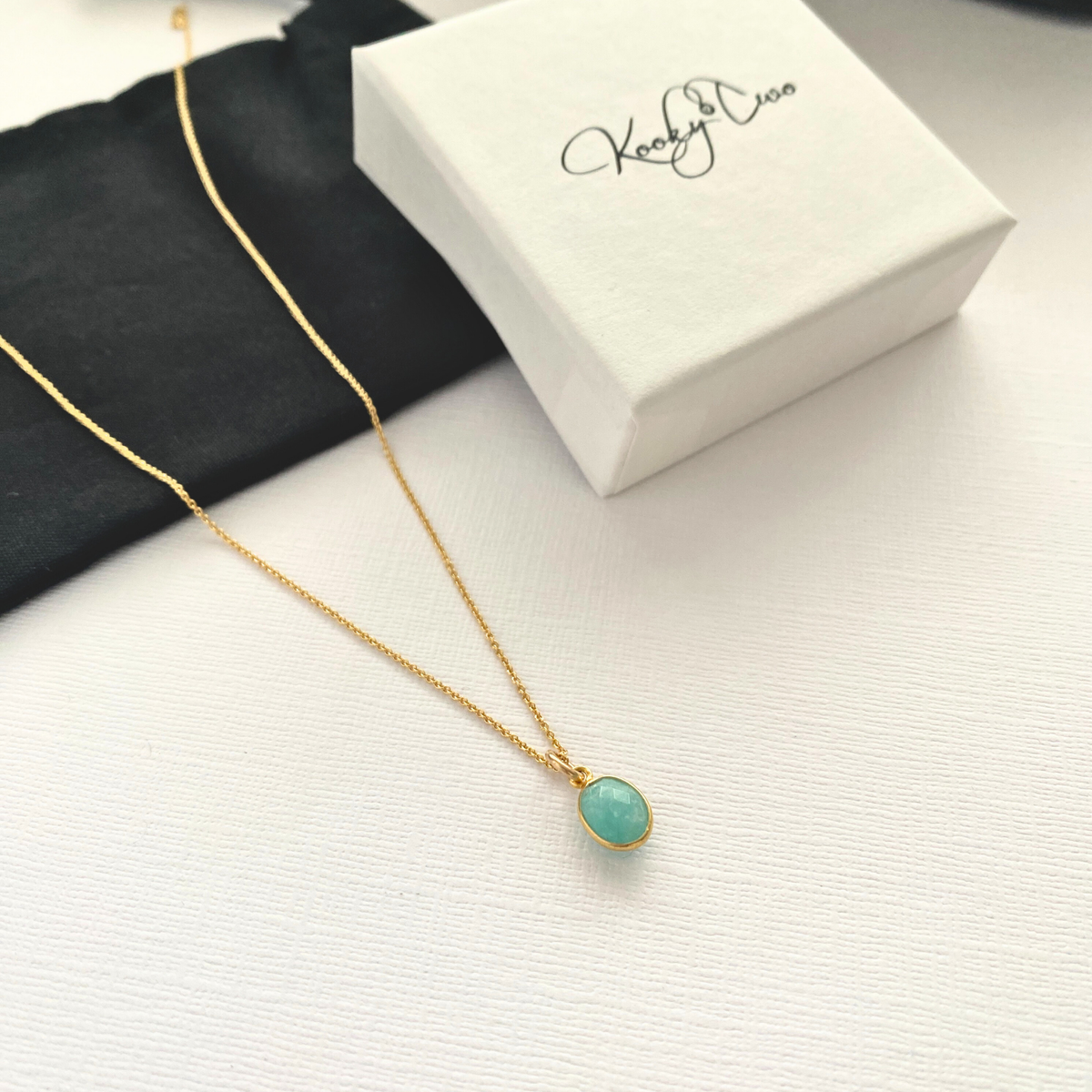 Gold necklace with amazonite pendant on a white surface next to a white gift box.