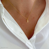 A close-up of a person wearing a delicate gold chain necklace with a small capital letter 'R' pendant, styled with a white collared shirt.