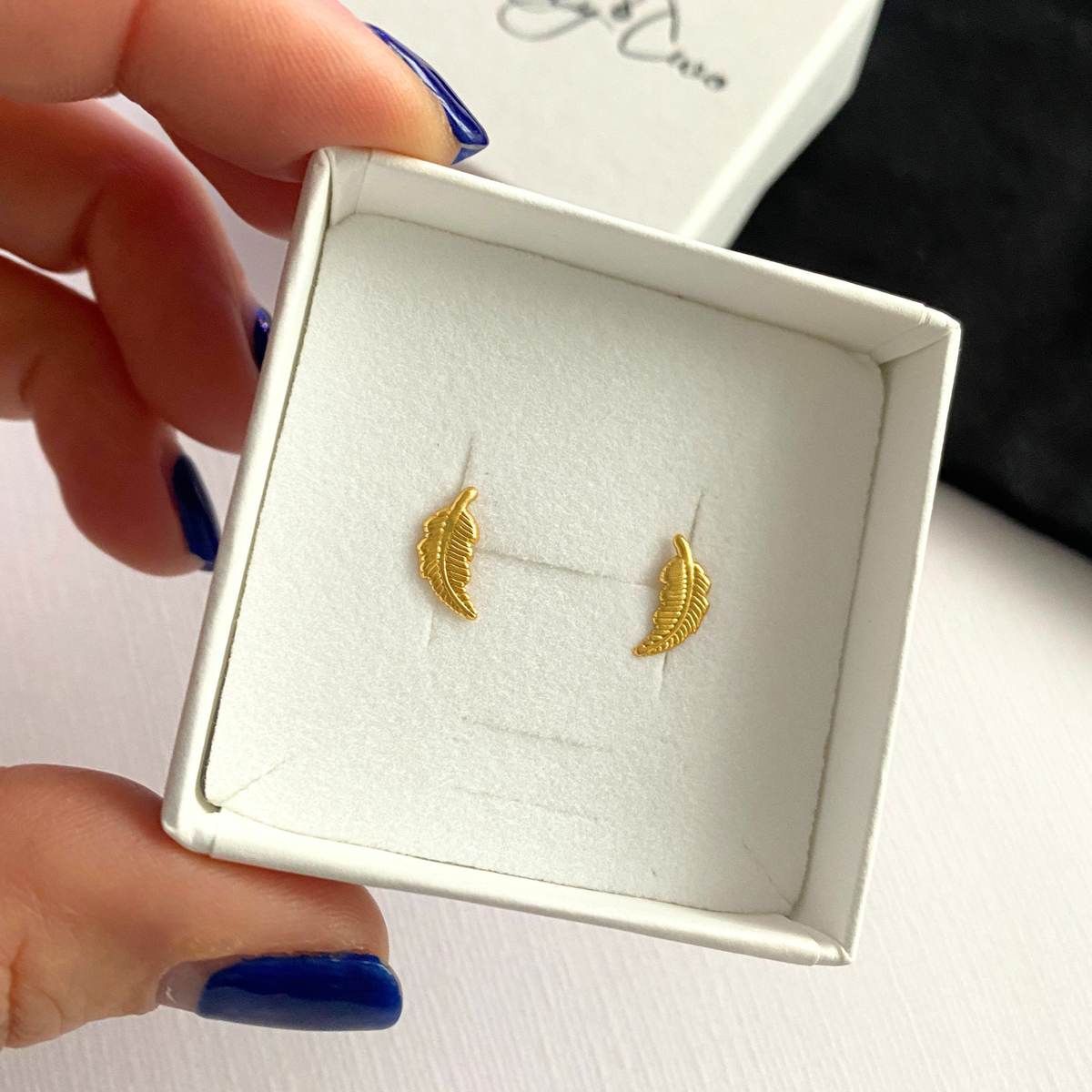 Gold feather-shaped earrings in a white jewellery box.