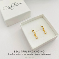 A pair of gold huggie hoop earrings with small crystal drops, presented in an open white jewelry box next to its branded white lid. Text at the bottom reads 'BEAUTIFUL PACKAGING' and 'Jewellery arrives in our signature box or stylish pouch'.