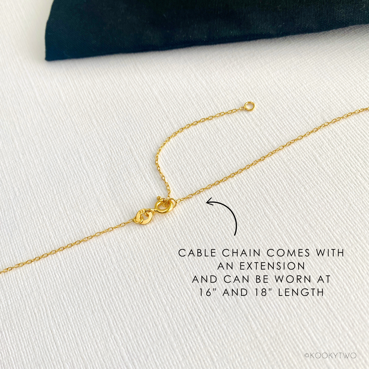 A close-up of a gold cable chain necklace with an extender clasp, shown on a textured white fabric background with a black velvet-like pouch. Text reads: 'CABLE CHAIN COMES WITH AN EXTENSION AND CAN BE WORN AT 16" AND 18" LENGTH'.