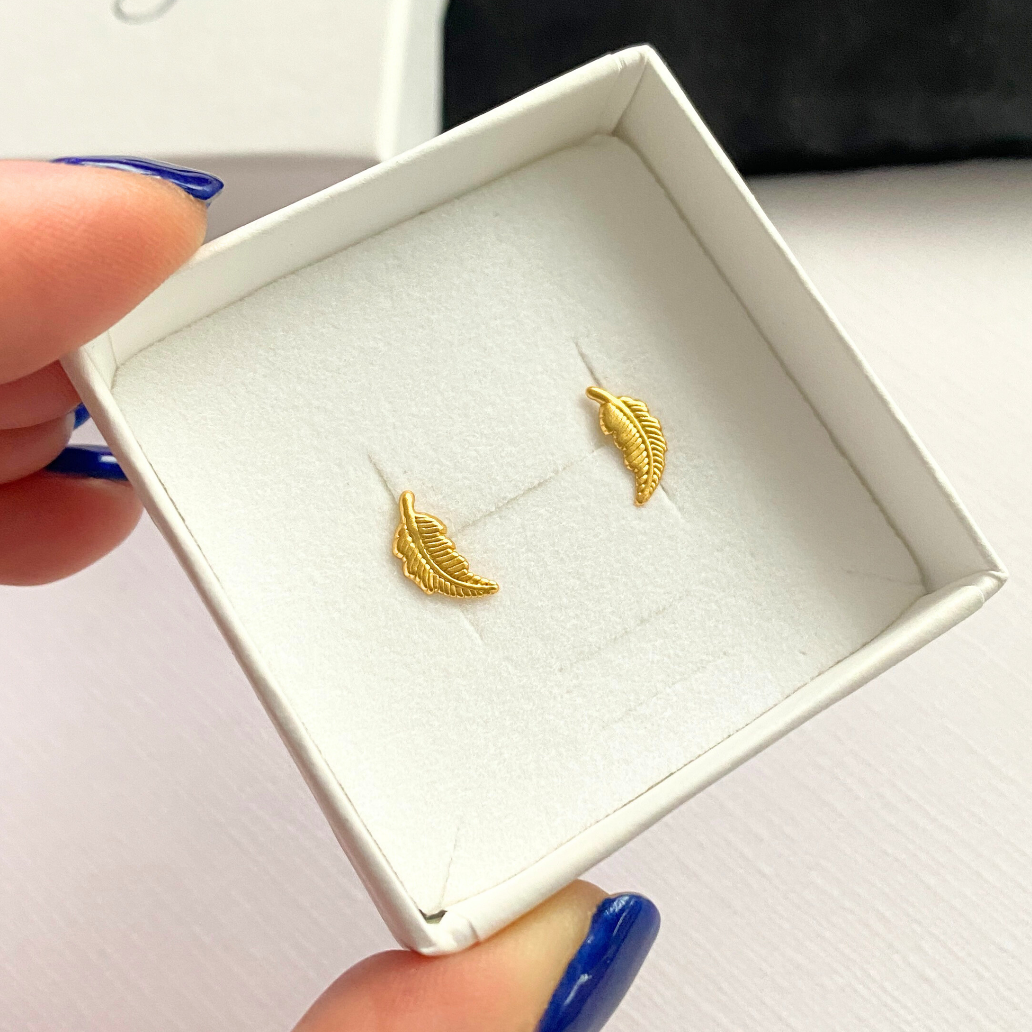 Gold feather-shaped earrings in a white gift box held by a hand.