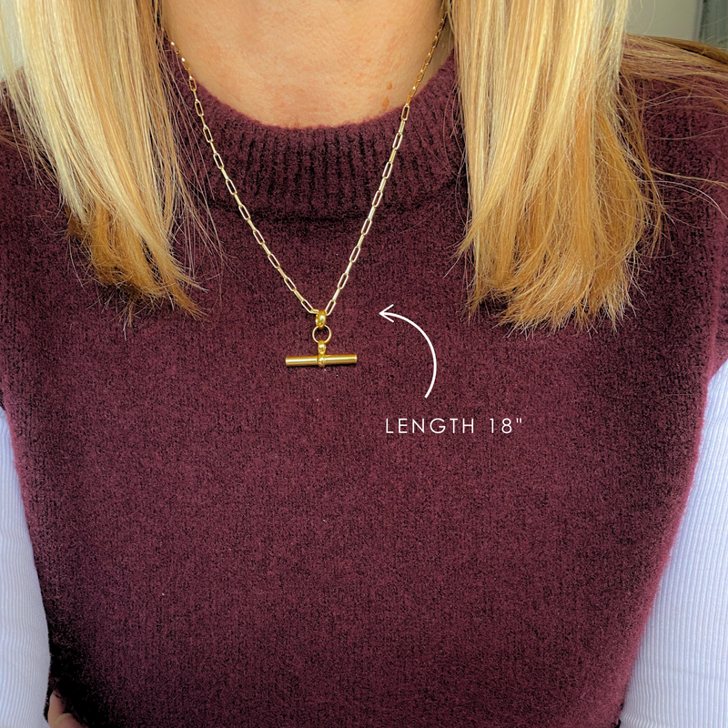 A bold and stylish eye-catching gold t bar necklace. A modern and elegant statement necklace, perfect for everyday wear. T Bar necklace shown worn over on-trend burgundy top.