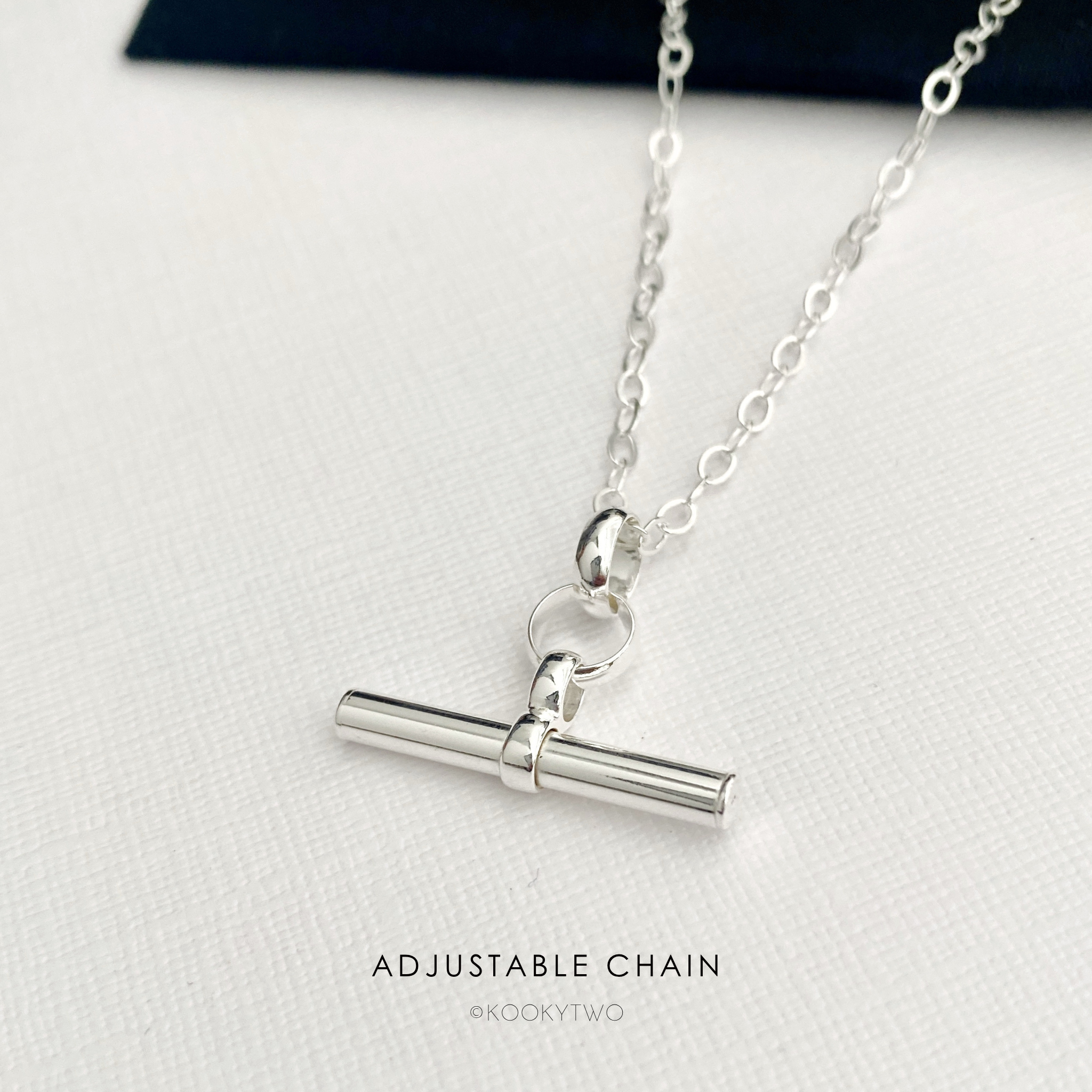 A silver adjustable t bar necklace on a textured white surface, with a dark navy velvet box corner visible in the background. The text 'ADJUSTABLE CHAIN' and the brand name 'KOOKYTWO' are printed at the bottom.