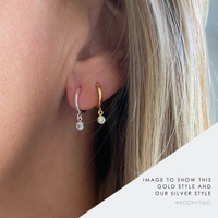 Close-up of a person's ear wearing two small huggie hoop earrings, one gold with a clear stone and one silver with a clear stone, with text overlay reading 'IMAGE TO SHOW THIS GOLD STYLE AND OUR SILVER STYLE' and 'KOOKYTWO'.