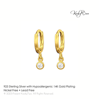 A pair of small gold huggie hoop earrings with a clear round gemstone dangle, shown on a white background with KookyTwo branding and product text.