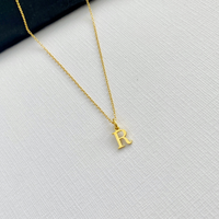 A delicate gold chain necklace with a small, polished gold initial pendant letter 'R', displayed on a textured white fabric surface with a dark corner accent.