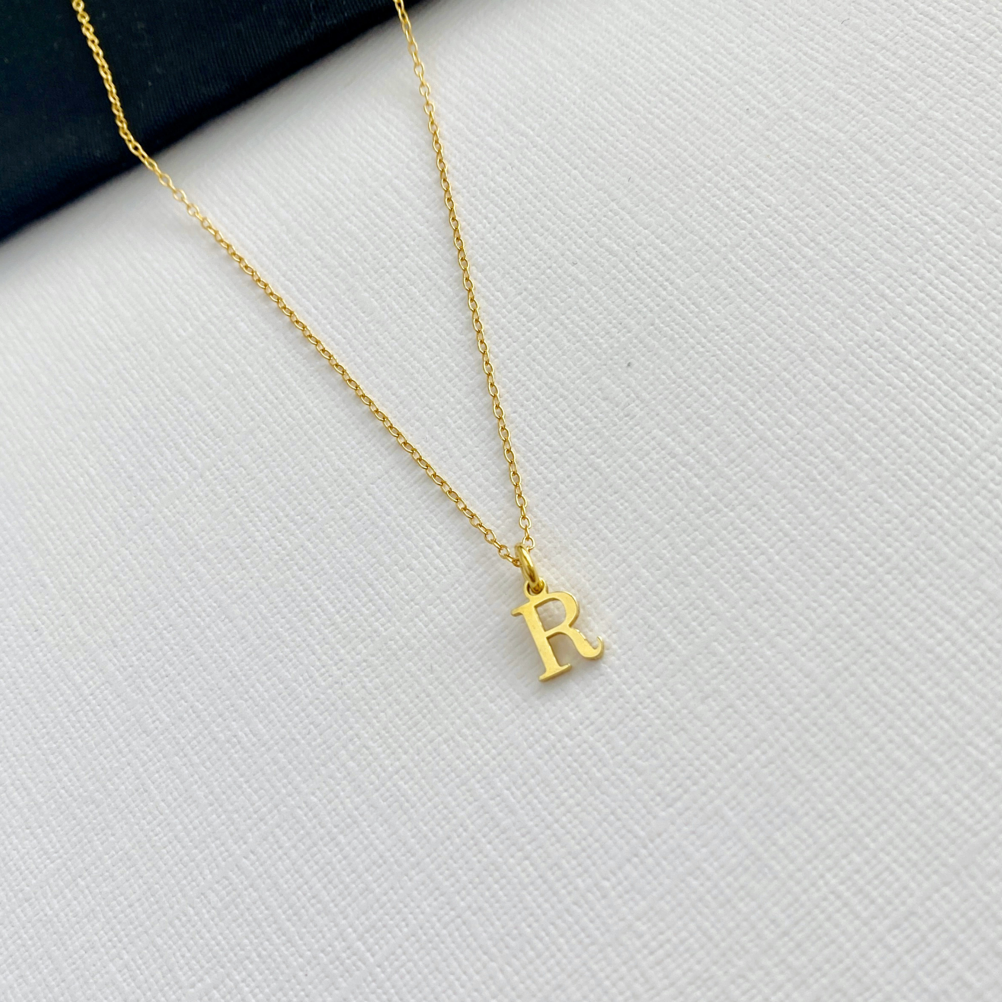 A delicate gold chain necklace with a small, polished gold initial pendant letter 'R', displayed on a textured white fabric surface with a dark corner accent.