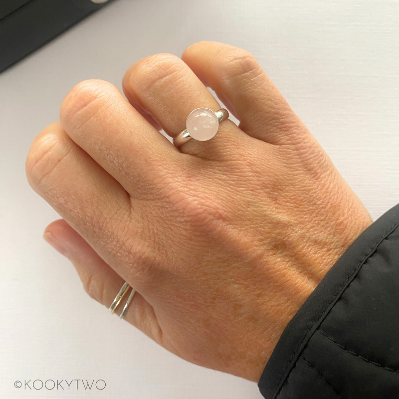 Lady wearing a ring with a pink gemstone, with a plain background. Gemstone is Rose Quartz on sterling silver adjustable ring base.