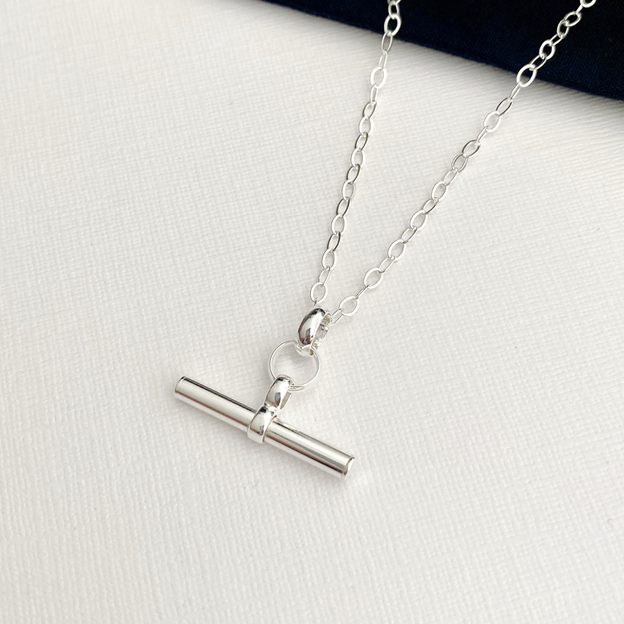 A silver necklace with a hanging bar pendant on a shiny oval trace chain, displayed on a textured white surface.