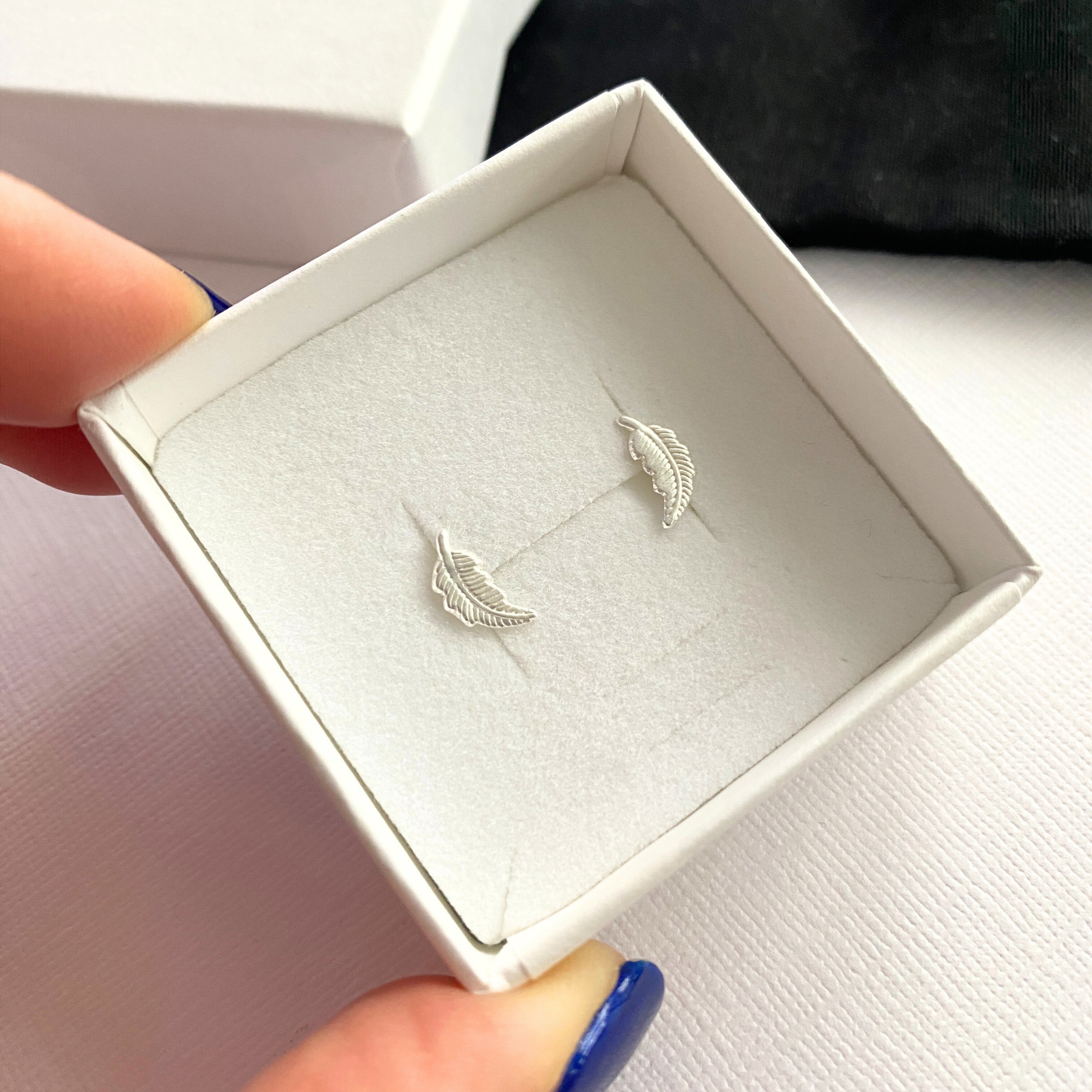 Silver feather earrings in a white box held by a hand with blue nail polish.
