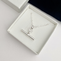 A silver t-bar pendant necklace displayed inside an open white gift box on a white surface.