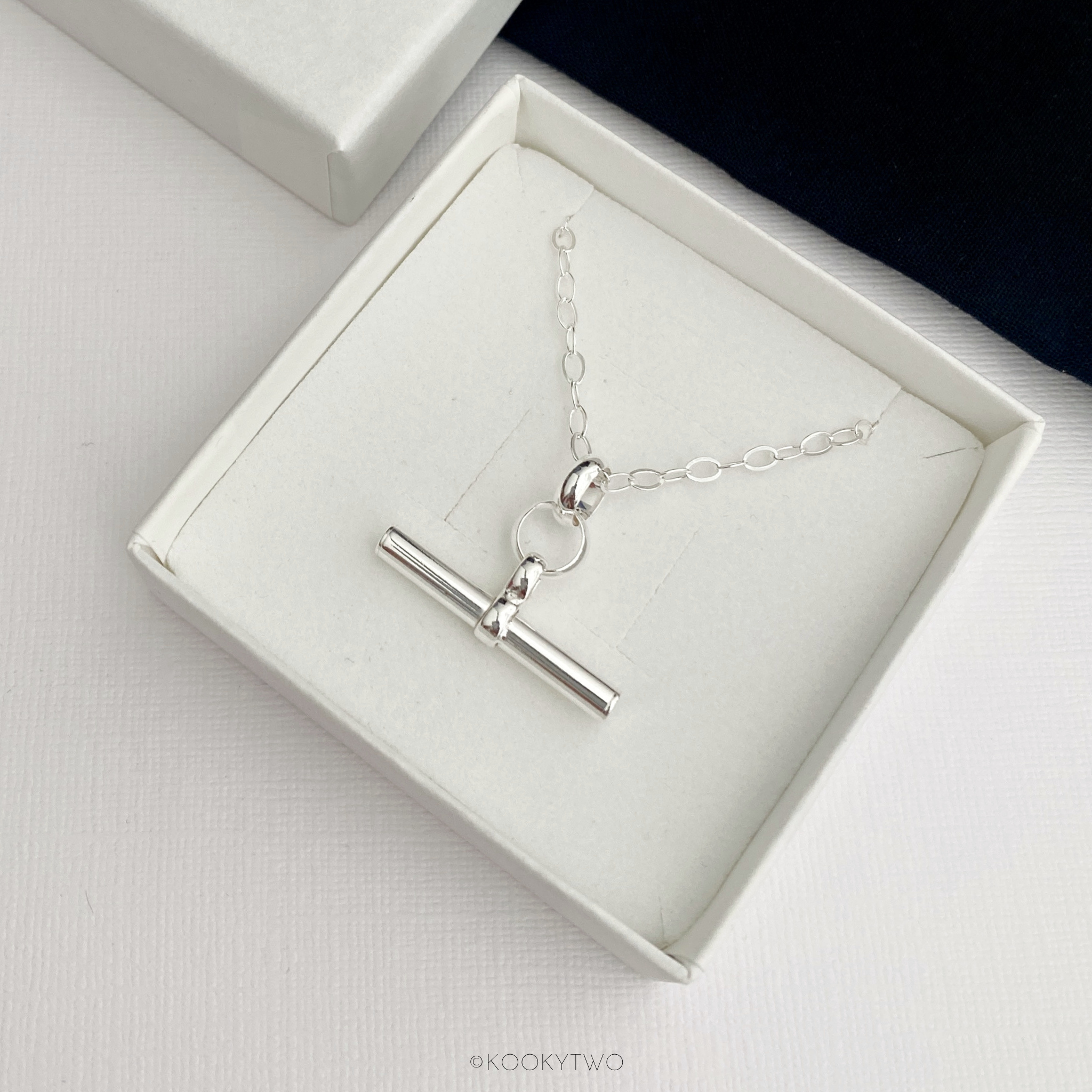 A silver hanging t bar necklace displayed inside an open white gift box, with a white textured surface.