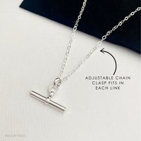 A silver adjustable chain necklace with a T-bar pendant, displayed on a textured white surface. A black arrow points to the chain with the text 'ADJUSTABLE CHAIN CLASP FITS IN EACH LINK'.
