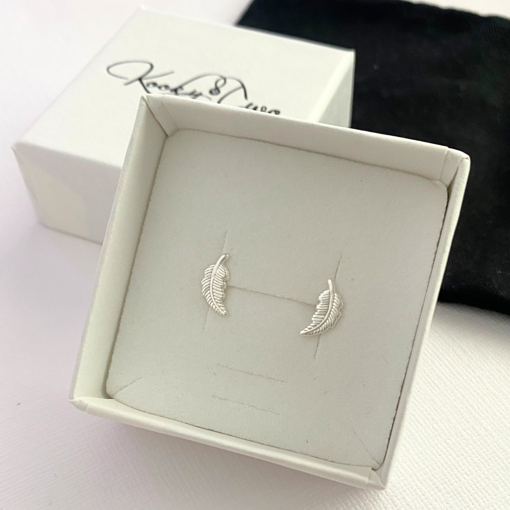 Silver feather-shaped earrings in a jewellery box with 'KookyTwo' branding.