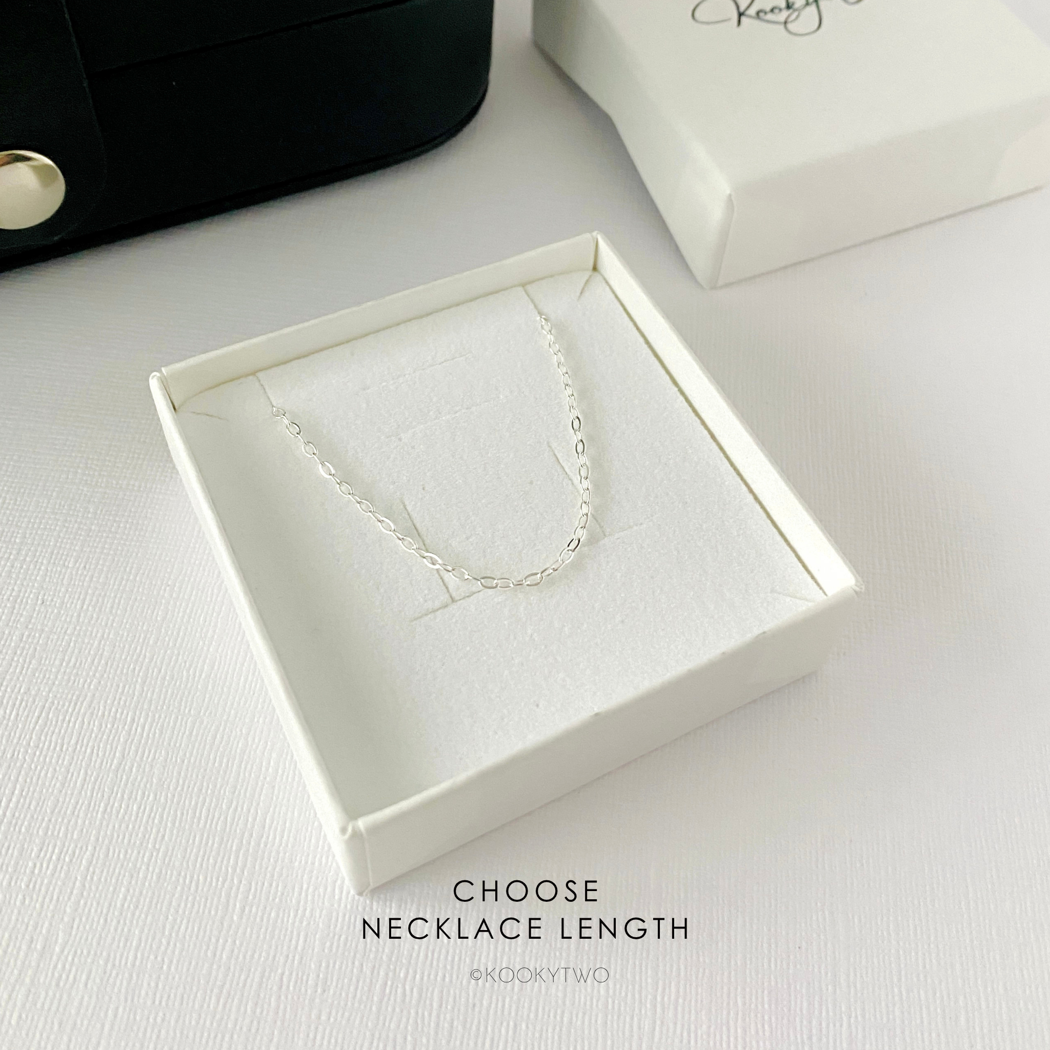 A delicate silver chain necklace displayed inside an open white jewellery gift box on a white surface, with a black jewellery box partially visible at the top left and a white box lid at the top right. Text at the bottom reads 'CHOSE NECKLACE LENGTH' and 'KOOKYTWO'.