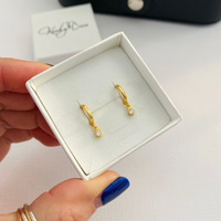 A close-up of a hand holding an open white jewelry box containing a pair of small gold huggie hoop earrings with clear gemstone drops. The background is a light white surface with a blurred KookyTwo brand box in the distance.