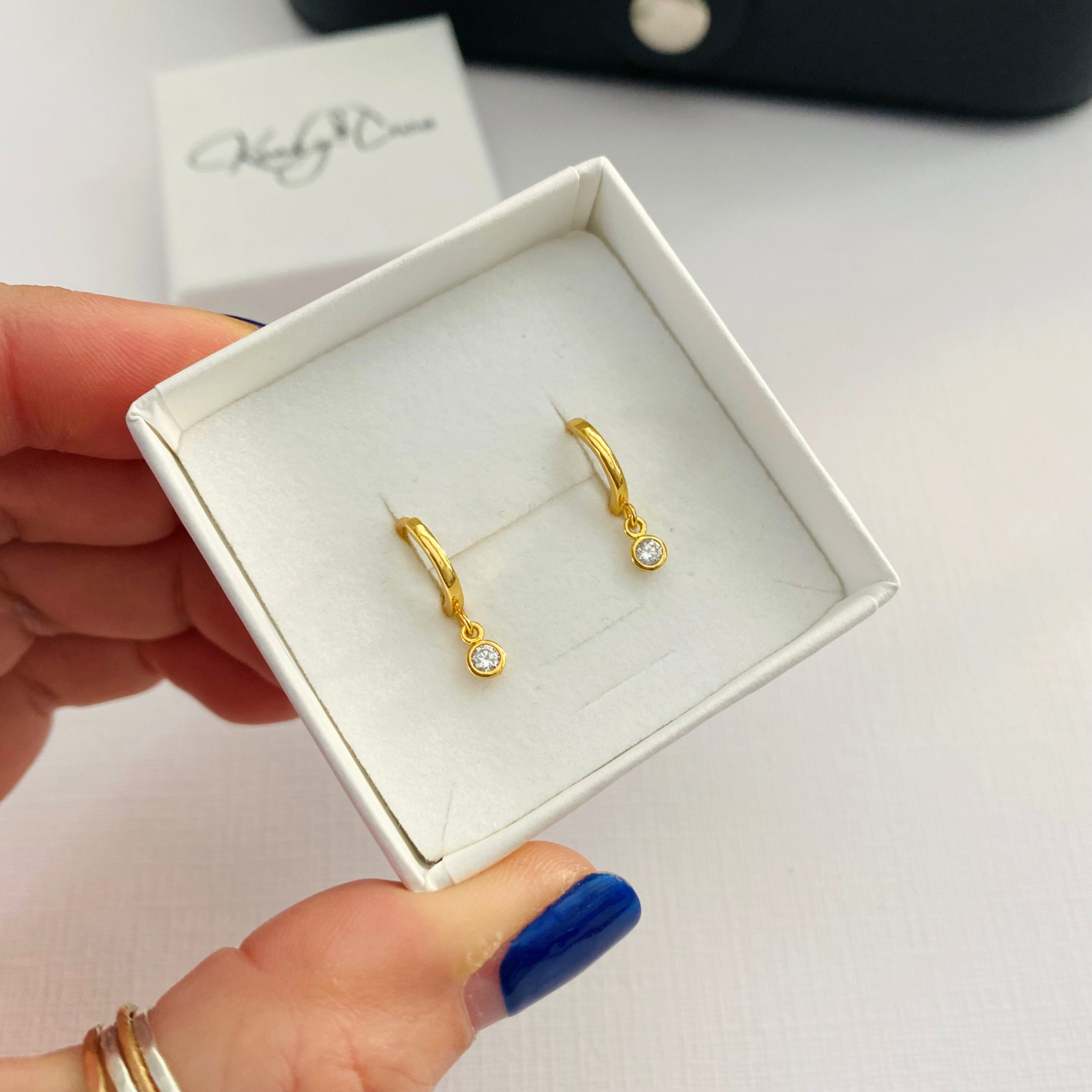 A close-up of a hand holding an open white jewelry box containing a pair of small gold huggie hoop earrings with clear gemstone drops. The background is a light white surface with a blurred KookyTwo brand box in the distance.