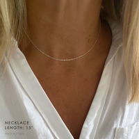 A close-up of a model's neck wearing a minimalist silver chain necklace. The model has blonde hair and is wearing a white shirt.