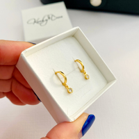 A pair of small gold huggie hoop earrings with clear round stones, displayed in a white jewelry box held by a hand with blue nail polish.