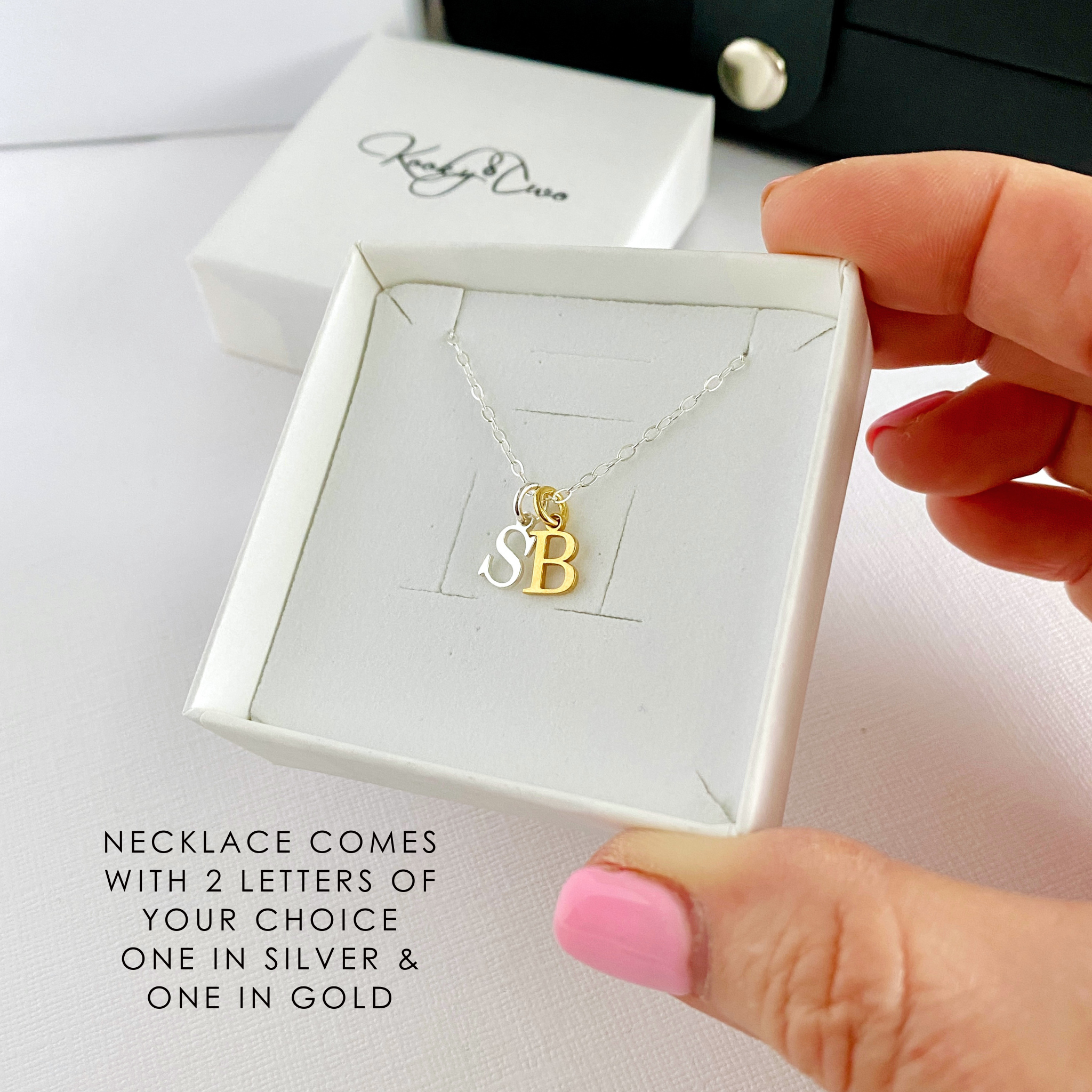 Gold and silver necklace with 'SB' letters in a white box, held by a hand.